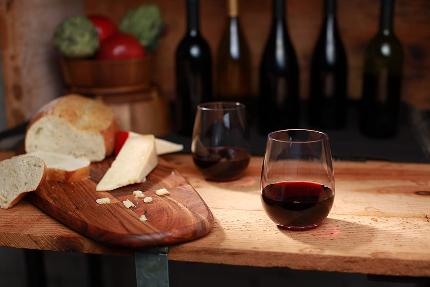 2 stemless unbreakable wine glasses with red wine in them with wine bottles in background and chopping board with bread.